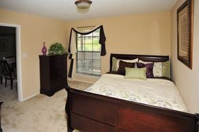 Overland Park apartment for rent - Bedroom with a dark wood bed and dresser, beige walls, carpeted floors, and natural light from a window.