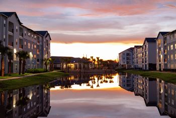 A serene view of apartment buildings reflected in a calm body of water during sunset.