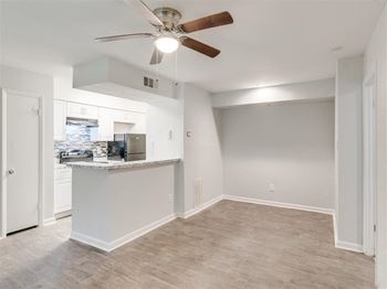 A room with a ceiling fan and a kitchen area in the background.