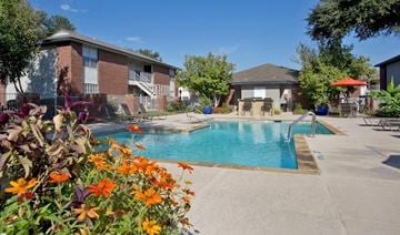 Burleson apartment for rent - A pool surrounded by flowers and chairs in front of a building.