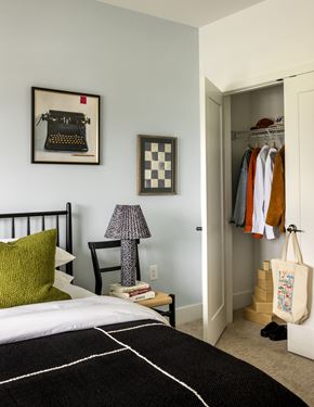 East Hanover apartment for rent - A bedroom with a bed, a chair, a lamp, a picture of a typewriter, a picture of a checkerboard, a wardrobe with clothes, and a tote bag on the floor.