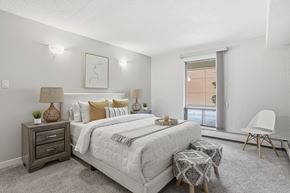 Edmonton apartment for rent - A bedroom with a large bed, a nightstand with a lamp, a chair, and a window with a view of a building outside.