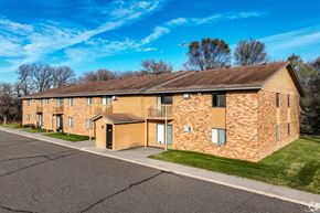 Elk River apartment for rent - A large brick building with a brown roof and a parking lot in front.