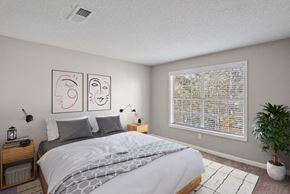 Charlotte apartment for rent - East Charlotte, NC, Affordable Apartments Near Albemarle Road Elementary - Woodland Estates - A Bedroom With A Large Bed, Wall Art, Wood Nightstands With Lamps, A Large Window, And A Potted Plant.