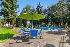 Federal Way apartment for rent - Federal Way, WA, Apartments Near Costco - Sunset Three Twenty - A Swimming Pool With Blue Lounge Chairs, Concrete Deck, Green Umbrella Seating Area, And Tall Evergreen Trees.