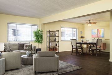 Long Beach apartment for rent - A living room with a grey couch, a coffee table, a dining table with chairs, a potted plant, and a shelf with books and decorations.