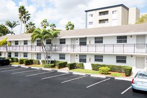 Hialeah apartment for rent - A parking lot in front of a white apartment building with a black car and a silver car parked.