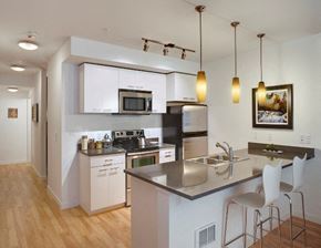 Seattle apartment for rent - One-Bedroom Apartments In Green Lake, Seattle, WA - Circa Green Lake - Kitchen With Hardwood Floor, Stainless Steel Appliances, Kitchen Bar, Pendant Lights, Quartz Countertops, And White Storage Cabinets.
