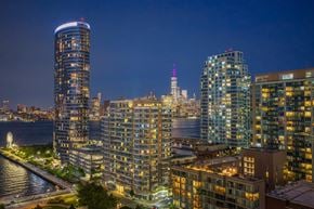 Jersey City apartment for rent - A cityscape at night with buildings illuminated and a body of water in the foreground.