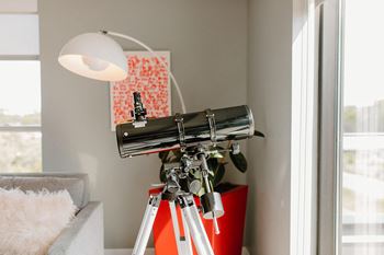 A telescope is on a tripod in a room with a white couch and a lamp.