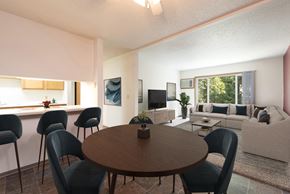 Bismarck apartment for rent - Bismarck, ND Bradbury Apartments. The room features a four-chair dining table with bar stools and a glimpse of the living room in the background. Soft lighting creates a warm and inviting atmosphere, while a window brings in natural light. It's a comfortable space for dining and relaxing with a seamless connection to the living room area