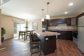 Bismarck townhouse for rent - Bismarck, ND Stonefield Townhomes. A kitchen with dark wood cabinets and a large island with a granite countertop. The space features a well-appointed dining table, modern kitchen appliances, and tasteful decor. Bright natural light fills the room from the glass sliding door.