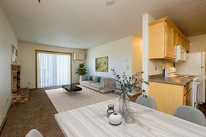 Fargo apartment for rent - Fargo, ND Windgate Apartments. a dining room with a living room in the background. the dining room table has four chairs. the living room has a couch, coffee table, and a glass sliding door.