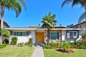 Downey multi-family for rent - A white house with a brown door and windows surrounded by greenery.
