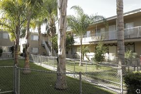 Bellflower multi-family for rent - A chain link fence surrounds a grassy area with palm trees and apartment buildings in the background.