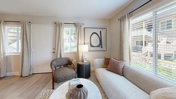 Poway apartment for rent - A living room with a grey armchair, a white sofa, a coffee table with a vase on it, and a large window with curtains.