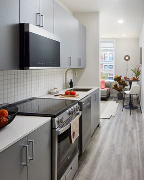Philadelphia apartment for rent - A photo of a galley kitchen looking towards the living room in the LVL West 1-Bedroom floorplan. The kitchen features gray cabinetry, a white backsplash, and light gray laminated flooring.