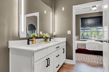 A bathroom with a vanity, mirror, and a bedroom in the background.