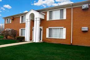 Romulus apartment for rent - Red brick apartment building with tall white pillar entrance with green maintained lawn and blue sky.