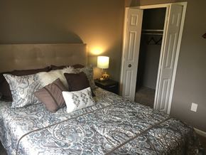 Brighton apartment for rent - Bedroom with tan walls and a tan headboard. The bed is made in a blue patterned bed spread and white and brow accent pillows. Next to the bed is a lamp on a side table. The white closets doors are open to the right of the bed.