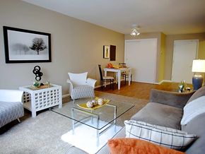 Columbus apartment for rent - Living area with brown couch on right wall with pattern pillows, glass coffee table in front, with two white chairs and end table on left wall.  Artwork above table in center.  In background is dining area and lamp.