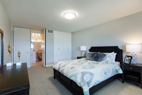 Detroit apartment for rent - Photo of a bedroom at The Jefferson. The walls are painted a light grey. There is a queen sized bed in the middle of the room with two wood-like black end tables on each side. The bed has a patterned comforter. There is a closet with two doors that pull open. There is light brown carpet throughout the room. Next to the closet there is an enterance to the bathroom.