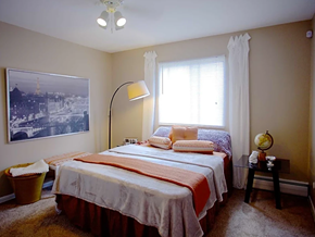 Columbus apartment for rent - Bed against window with white curtains on each side of window.  Two-tone beige walls with white accent trim.  Striped orange bedding, beige carpet and photo on left wall.  Basket under photo and table to right of bedside.