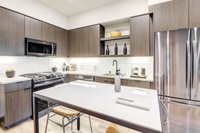 Oakland apartment for rent - Downtown Oakland, CA, Luxury Apartments for Rent - Edson House - Modern Kitchen With Wood Tone Cabinets, Stainless Steel Refrigerator, White Tile Backsplash, And A Large Island Table.