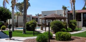 Tucson apartment for rent - A gazebo is surrounded by bushes and palm trees in front of a building.