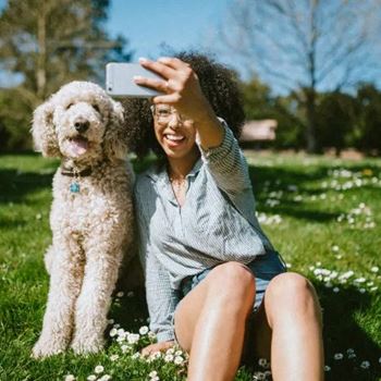 A woman sitting in the grass with her dog taking a picture with her cell phone