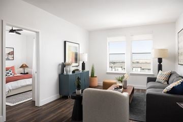 Denver apartment for rent - A living room with a grey couch, a grey armchair, a small table with a plant on it, and a bed with a red blanket in the background.