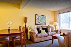 Kerman apartment for rent - A living room carpet, a yellow accent wall, and a window with blinds. The room is staged with a couch, coffee table, a painting, and a dining room table set for 4.
