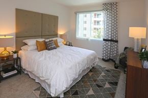 Anaheim apartment for rent - A bedroom with light colored carpet, white walls, and a window. The room is staged with a bed, nightstands, and a dresser.