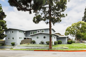 Inglewood apartment for rent - A modern, multi-story gray apartment building surrounded by greenery. The structure features large windows and a landscaped yard with shrubs. Tall trees provide shade in the vicinity, and a gray sidewalk runs along the front. The sky is partly cloudy, creating a serene atmosphere.