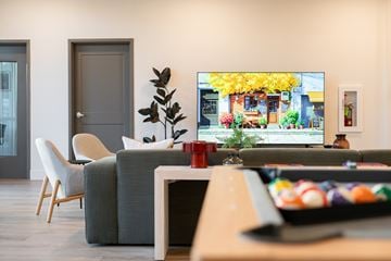 Garden City apartment for rent - A living room with a grey sofa, a white chair, a wooden table, and a television displaying a yellow building.