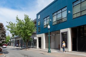 Bremerton apartment for rent - A woman in a straw hat walks down a sidewalk past a blue building at B Flats Apartments, Bremerton, 98337