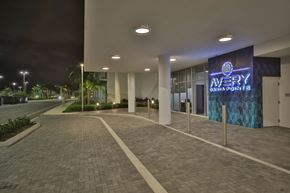 Dania Beach apartment for rent - The entrance to this apartment building features a modern, well-lit design with sleek columns, a paved walkway, and an illuminated sign that reads "Avery Danna Pointe" in blue neon. The surroundings are lush with greenery, and the area has a clean, contemporary look, illuminated by overhead lighting.