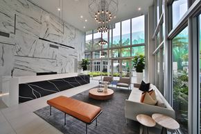 Dania Beach apartment for rent - Lobby with a stunning marble front desk with a black and white design, seamlessly integrated into the space. The modern aesthetic is complemented by sleek marble walls, large windows allowing abundant natural light, and a stylish seating area with a leather bench and contemporary furniture. Potted plants and unique lighting fixtures further elevate the sophisticated setting.