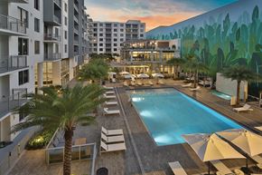 Dania Beach apartment for rent - Resort style pool within the community, with balconies overlooking it, a large nature inspired mural, a marble waterfall at the spa, cabanas, palm trees scattered around, and white pool lounge chairs.