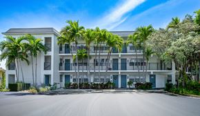 Boca Raton apartment for rent - A white apartment building with blue doors and windows is surrounded by palm trees.
