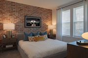 Antioch apartment for rent - Dog-Friendly Luxury Apartments In Antioch, TN - Mainstead At Century Farms - Warm Bedroom Featuring A Red Brick Accent Wall, A Grey Upholstered Bed, And Industrial-Style Gold Lamps.