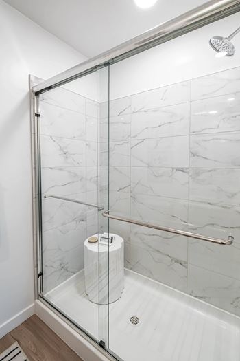 A white marble tiled shower with a glass door and a toilet paper roll on the floor