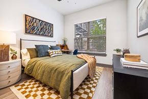 Duluth apartment for rent - A bedroom with a bed, a dresser, a window with blinds, a painting on the wall, a plant on the nightstand, and a rug on the floor.