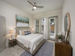 Jacksonville apartment for rent - One-Bedroom Luxury Apartments in Jacksonville, FL - Terrabella - Bedroom with Carpeted Flooring, Large Bed, Nightstands with Lamps, Wooden Dresser, Round Mounted Mirror, White Walls, Ceiling Fan, Window, and Glass Door with Access to Patio.