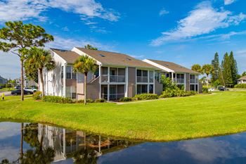 A building with a grey roof and white walls is surrounded by greenery and a body of water