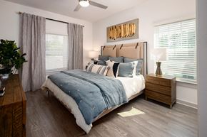 Ridgeville apartment for rent - Pet-Friendly Luxury Apartments in Ridgeville, SC - Preserve at Ridgeville - Bedroom With Upholstered Bed, Blue Bedding, Wooden Nightstand With Lamp, Two Large Windows With Curtains And Blinds.