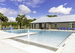 Titusville apartment for rent - One-Bedroom Apartments in Titusville, FL - Riverwalk Grand - Resort Style Pool With Shallow Water Lounge Area, Splash Fountain, Blue Chairs, Palm Trees, And Clubhouse Building.
