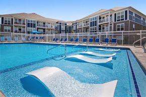 Holland apartment for rent - Holland, MI, Pet-Friendly Apartments - Shoreline Flats - Resort-Style Swimming Pool with In-Water Loungers, Surrounded by a White Fence, Blue Sun Loungers, and Shaded Lounge Chairs, Overlooking the Apartments.