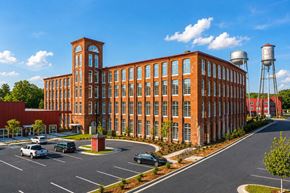 Warrenville apartment for rent - Upscale Apartments in Aiken, SC - Warren Mill Lofts - Historic Brick Apartment Building Exterior With Tall Arched Windows, Landscaped Grounds, Parking Areas, And Two Water Towers Under A Blue Sky.