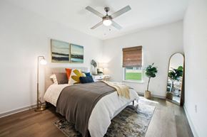 Flagler Beach apartment for rent - Luxury Apartments in Flagler Beach, FL, with In-Unit Washer and Dryer - Preserve at Flagler Beach - Bedroom with Ceiling Fan, Bed, Area Rug, Wood-Inspired Flooring, High Ceiling, Desk with Lamp and Chair, and Window.